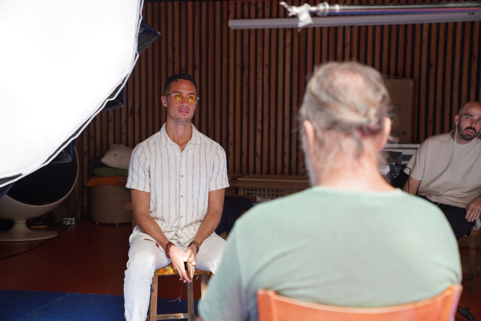 Jonathan Ramalho sits on a stool facing a man seen from behind, with a large softbox light and a crew member in the background of a wood-panelled room.