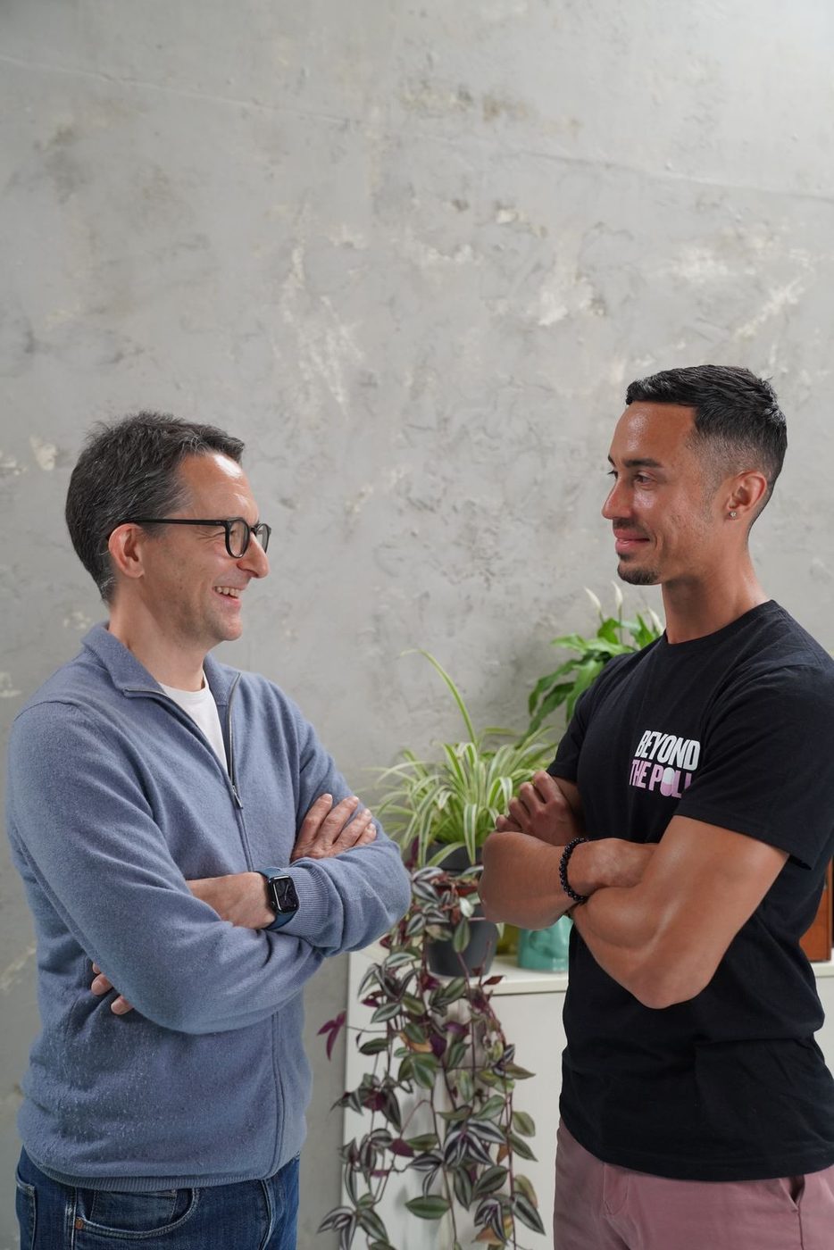 Jonathan Wolf and Jonathan Ramalho facing each other with arms crossed, smiling, in front of a pale concrete wall and a shelf of trailing plants.