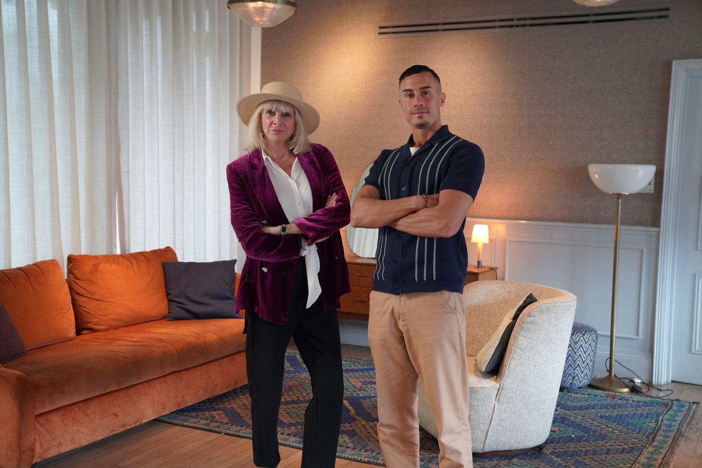 Marisa Peer, in a wide-brimmed hat and burgundy velvet jacket, stands beside Jonathan Ramalho, whose arms are crossed, in a living room with an orange sofa.