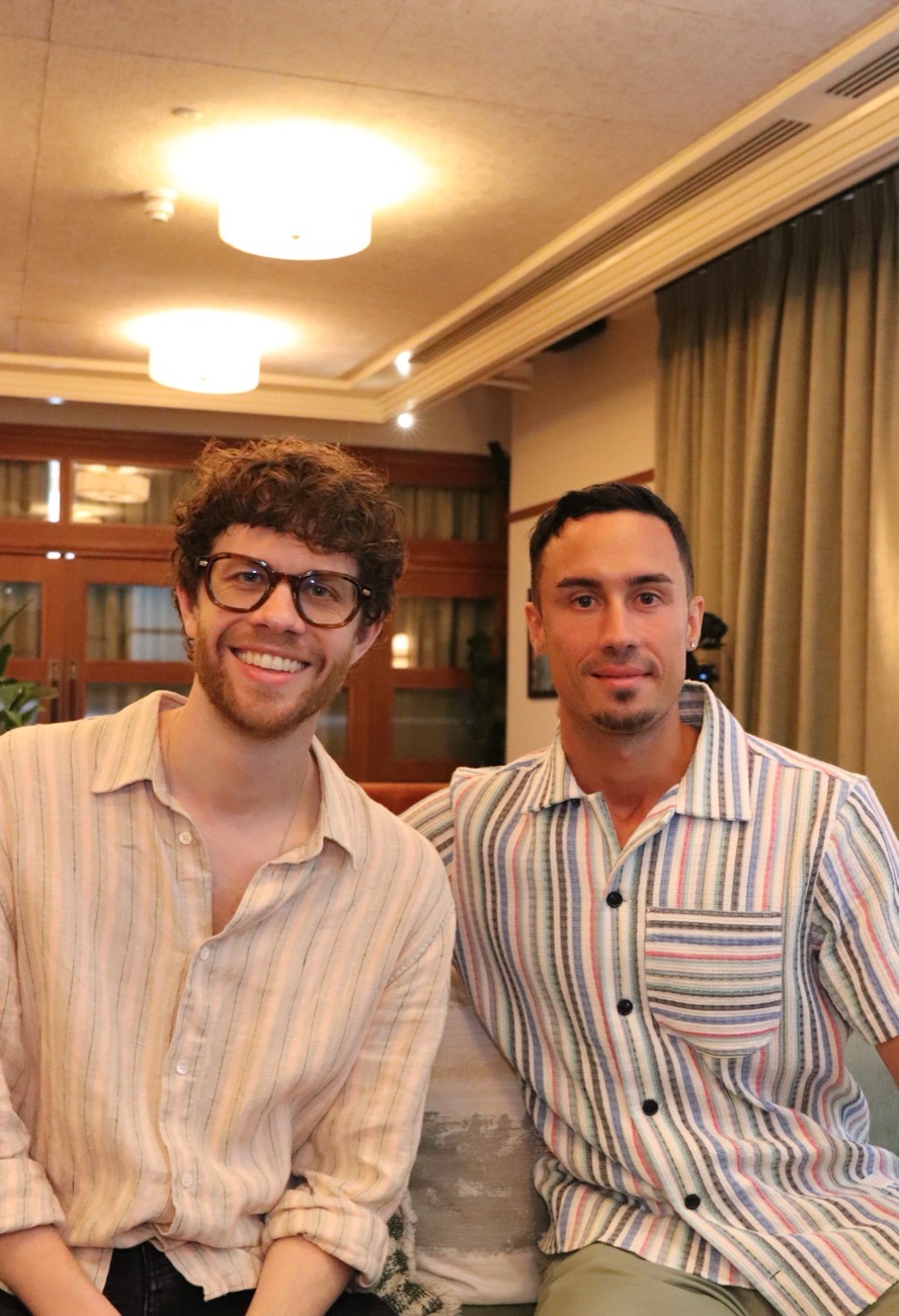 Dr Simon Ruffell and Jonathan Ramalho sitting side by side on a sofa in a warmly lit lounge, both in striped shirts.