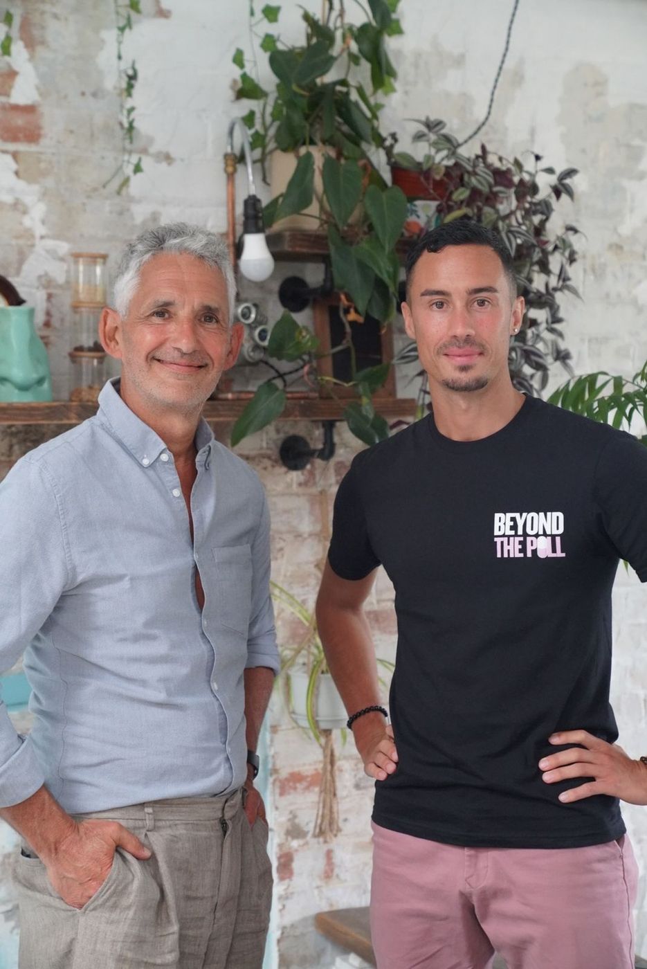 Tim Spector and Jonathan Ramalho standing together in front of a whitewashed brick wall hung with plants; Jonathan wears a Beyond The Pill T-shirt.