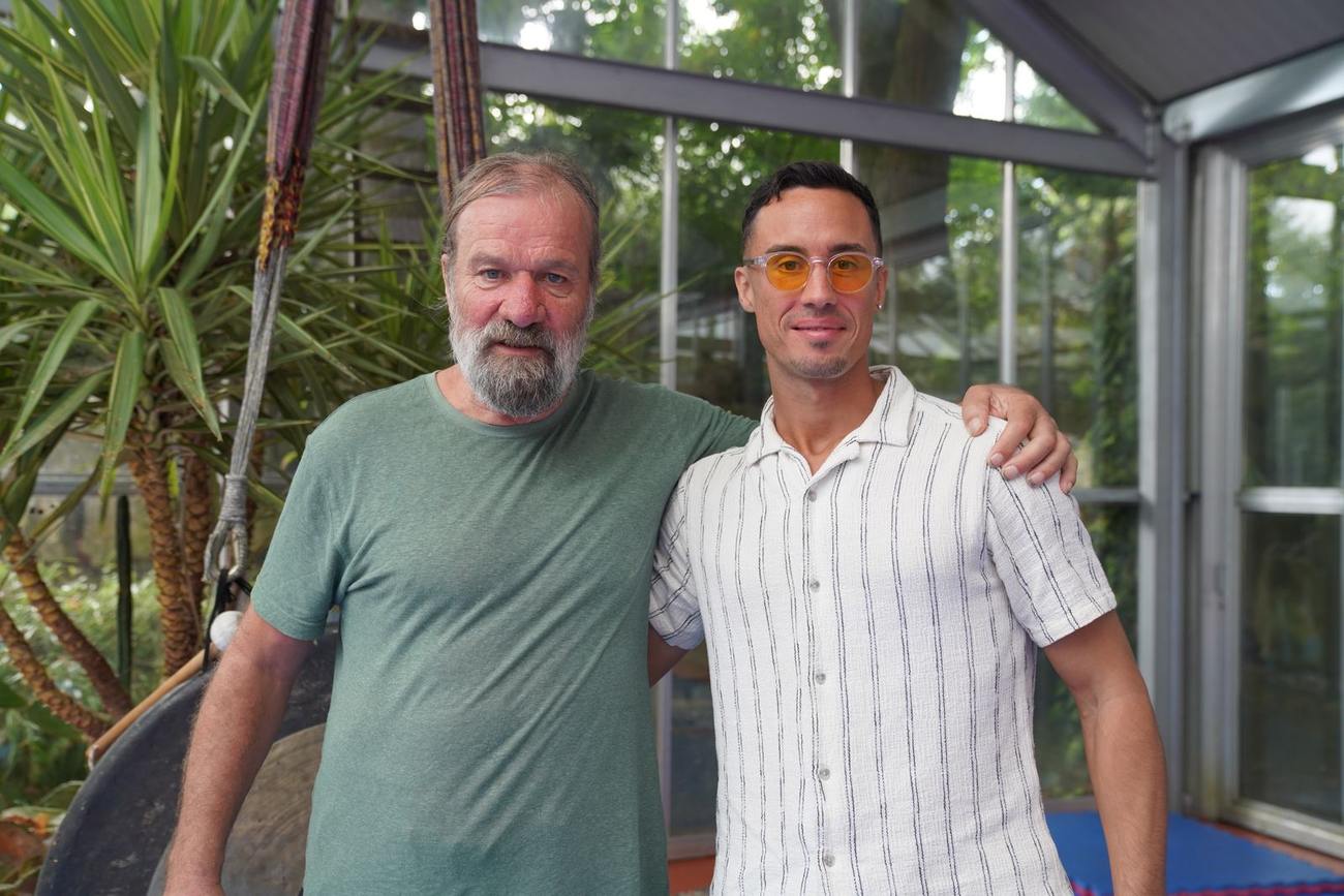 Wim Hof and Jonathan Ramalho standing side by side, arms around each other, in a glasshouse with palms and blue floor mats.