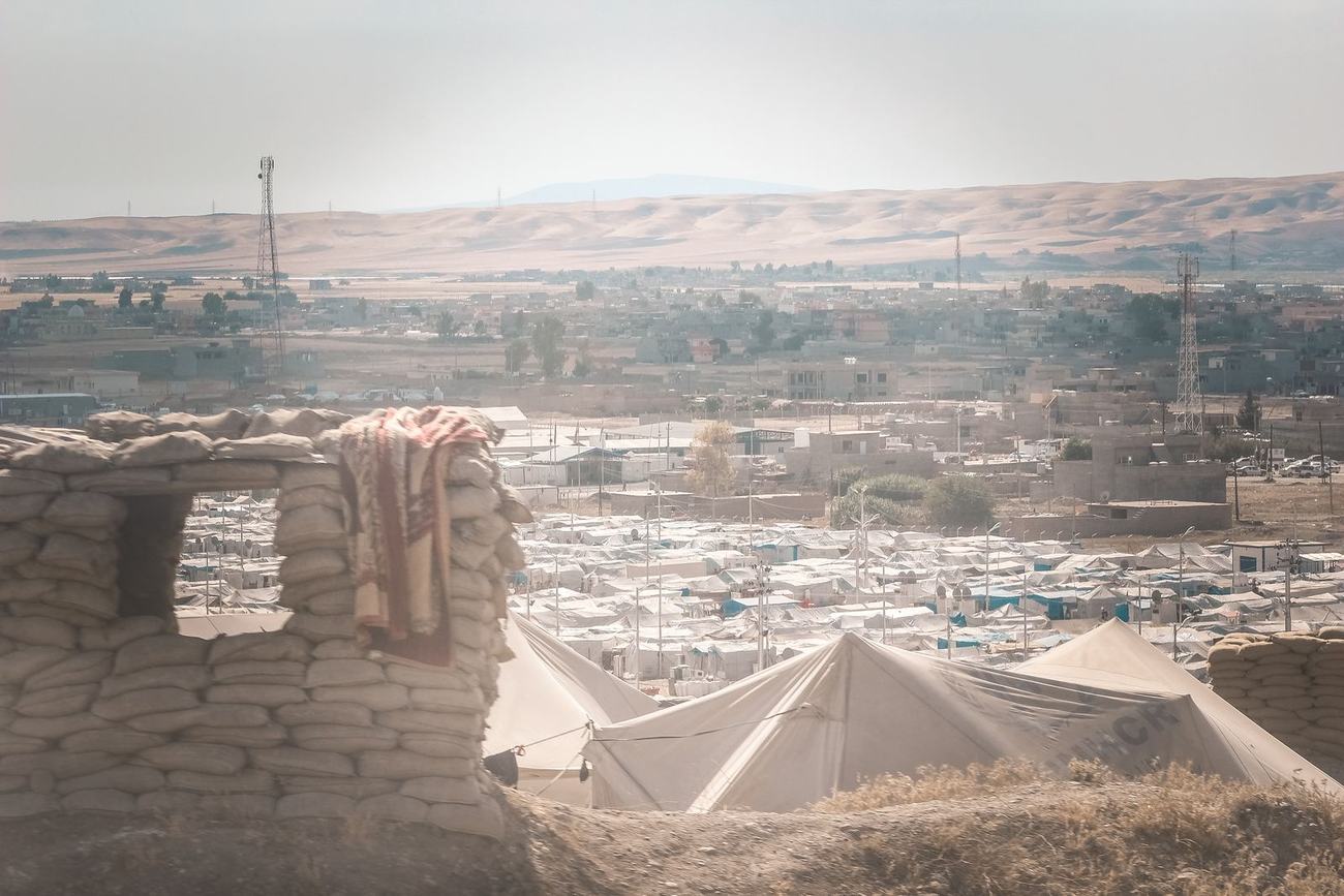 View from behind a sandbag wall over a large camp of white tents and low buildings, with dry hills on the horizon.