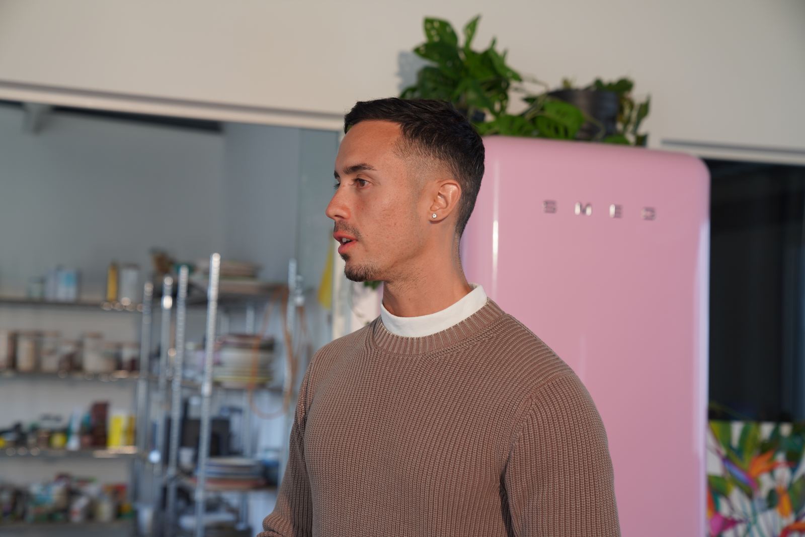 Jonathan Ramalho in a brown knit sweater, looking to the side, in a kitchen with a pink refrigerator and open shelving.