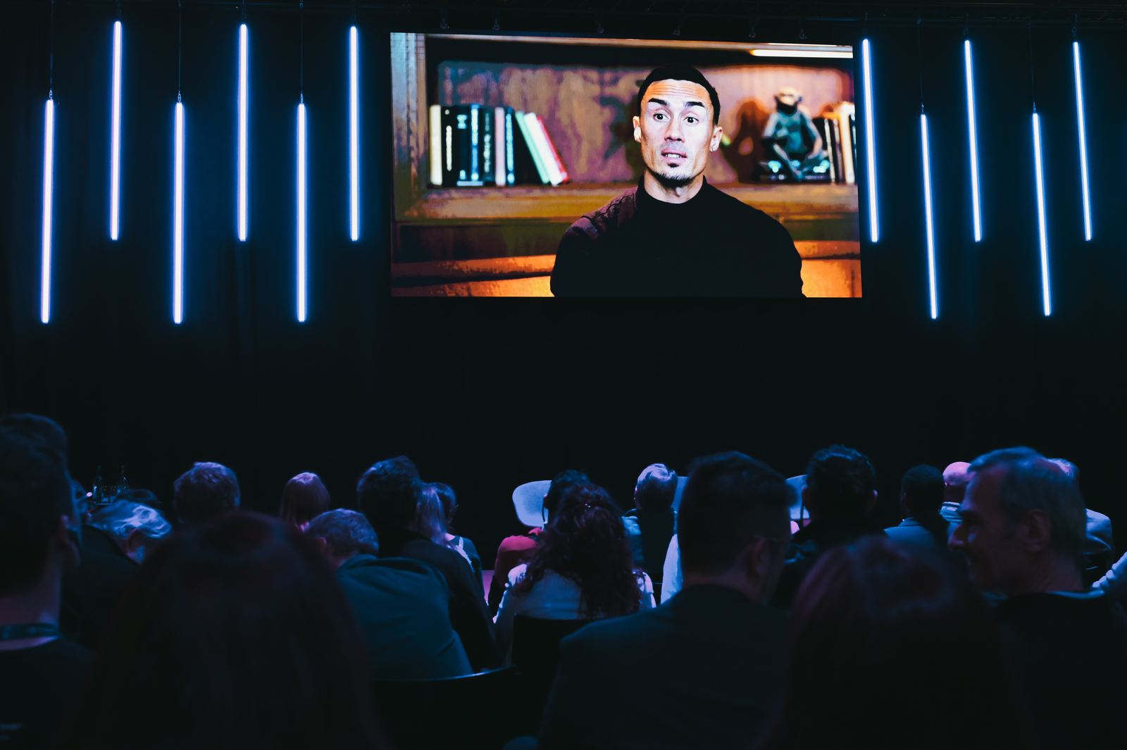 An audience in a dark hall watches a large screen showing Jonathan Ramalho speaking in front of a bookshelf, framed by vertical blue light bars.