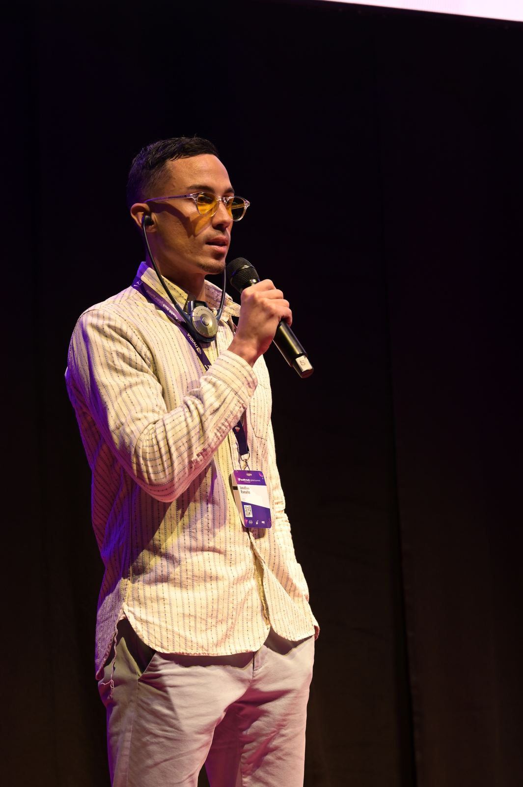 Jonathan Ramalho speaking into a handheld microphone on a dark stage, wearing a striped shirt, amber glasses, headphones around his neck and a conference lanyard.